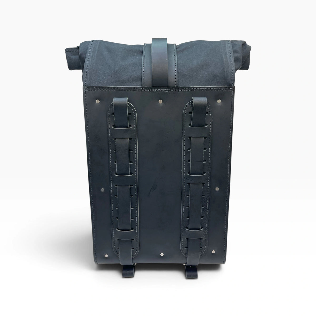 A black leather and canvas utility backpack shown from the back, featuring two vertical leather strap panels with metal rivets and a top handle, set against a plain white background.